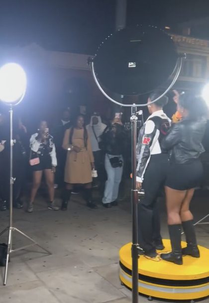 Two performers on the yellow booth at a night session, lit by studio lights, with a crowd watching and filming on their phones
