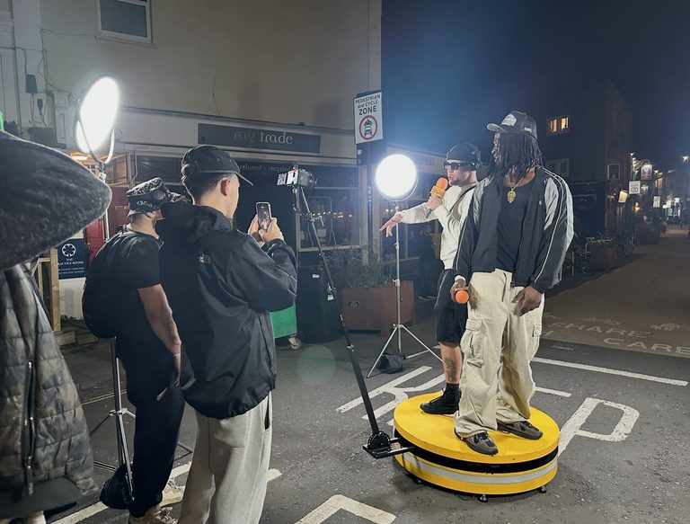 Two artists performing on the yellow On Da Booth platform at night, holding On Da Booth microphones, with studio lights, a camera on a boom arm and people filming on their phones