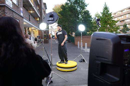 A London street session at golden hour: an artist on the yellow booth between ring lights, with the crowd and a speaker in the foreground